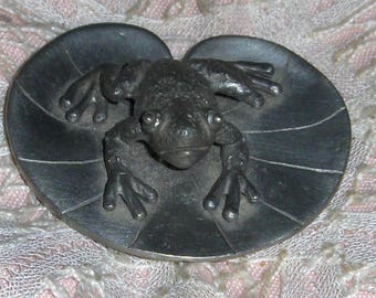 May include: A pewter frog figurine resting on a lily pad. The frog is detailed with textured skin and prominent eyes. The lily pad has veining details. The figurine is a decorative item.