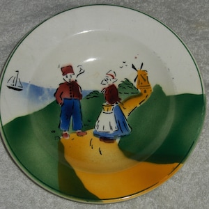 May include: A vintage ceramic plate with a green, yellow, and blue landscape scene. The scene features two figures, a windmill, and a sailboat.