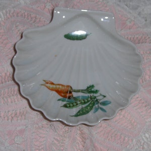 K&ouml;nnte beinhalten: Eine wei&szlig;e Porzellanschale in Muschelform mit einem gr&uuml;nen Blatt, einer orangefarbenen Karotte und gr&uuml;nen Erbsen, die darauf gemalt sind.