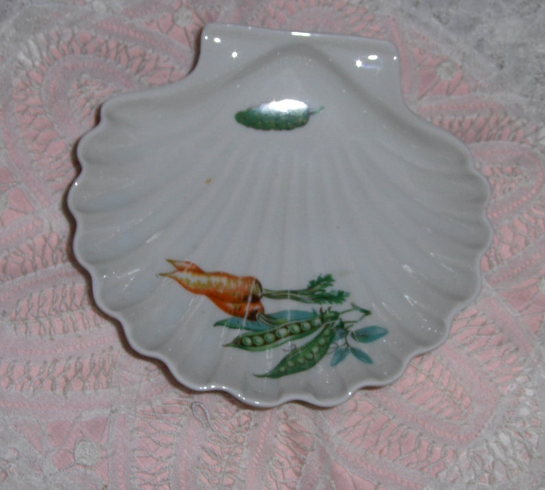 K&ouml;nnte beinhalten: Eine wei&szlig;e Porzellanschale in Muschelform mit einem gr&uuml;nen Blatt, einer orangefarbenen Karotte und gr&uuml;nen Erbsen, die darauf gemalt sind.