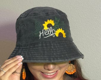 May include: Black bucket hat with embroidered yellow sunflowers and the word "Hello" in white script. The hat has a wide brim and is made of a soft, textured fabric. The hat is a casual accessory.