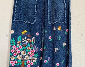 May include: Dark blue denim pants with embroidered floral and insect designs. The pants have two back pockets with frayed edges and elastic cuffs. The floral embroidery includes pink, white, and yellow flowers, with green leaves and brown stems.