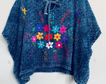 May include: A denim-style top with colorful floral embroidery. The top features a tie-up neckline and short sleeves. The embroidered flowers are in shades of pink, yellow, blue, and red, creating a vibrant contrast against the blue fabric.