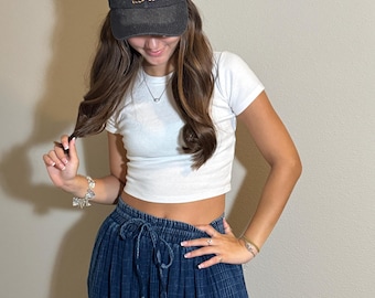 May include: A dark gray baseball cap with the word "Ashes" embroidered in orange, pink, and green. The cap is worn by a person wearing a white crop top and a blue skirt.