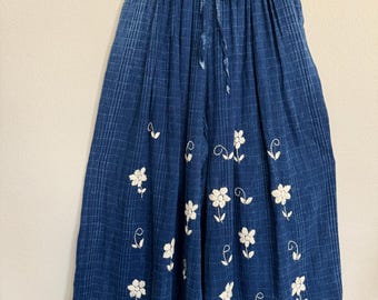 May include: Blue denim culottes with a white floral embroidery pattern. The culottes have a drawstring waist and are hanging on a black hanger. The fabric has a subtle plaid design.