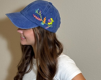 May include: A blue baseball cap with embroidered floral design in shades of pink, yellow, orange, and purple. The cap has a curved brim and a fabric strap closure. The cap is made of a soft, washed cotton material.
