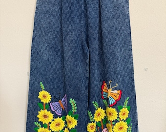 May include: Blue denim pants with an elastic waistband. The pants feature embroidered sunflowers and butterflies in yellow, purple, orange, and pink. The embroidery is detailed and adds a decorative element to the casual pants.