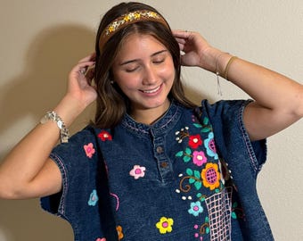 May include: A denim top with colorful floral embroidery and a brown headband with floral accents. The top has a button-up neckline and short sleeves. The embroidery includes pink, red, yellow, and blue flowers.