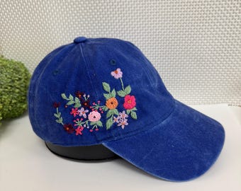 May include: A blue baseball cap with embroidered floral design. The flowers are in shades of pink, orange, and purple with green leaves. The cap has a curved brim and a button on top.
