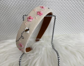 May include: A beige headband with embroidered pink flowers and a bee. The headband has a brown interior and is displayed on a silver metal stand. The flowers are in full bloom, and the bee is in flight.