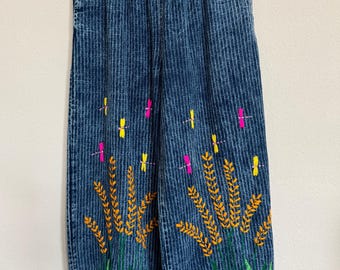 May include: Blue denim pants with vertical stripes and an elastic waistband. The pants feature embroidered wheat stalks in orange and green, with yellow and pink dragonfly accents. A casual, comfortable style.