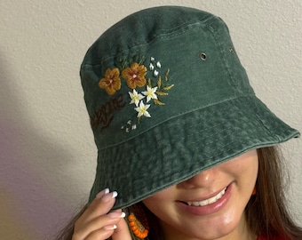 May include: Green bucket hat with embroidered floral design in shades of orange, white, and brown. The word "explore" is embroidered above the flowers. The hat has a classic, casual style.
