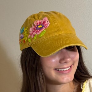 Puede incluir: Gorra de béisbol de color amarillo mostaza con diseño floral bordado. Las flores incluyen flores rosas, moradas y azules con hojas verdes. La gorra tiene una visera curva y un diseño clásico de seis paneles.