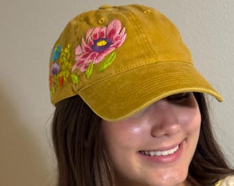 May include: Mustard yellow baseball cap with embroidered floral design. The flowers include pink, purple, and blue blooms with green leaves. The cap has a curved brim and a classic six-panel design.