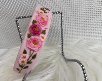 May include: A pink headband with embroidered pink and yellow flowers and green leaves. The headband is displayed on a silver metal stand against a white, fluffy background. The floral embroidery adds a delicate touch.