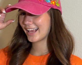 May include: A fuchsia baseball cap with embroidered floral designs in shades of blue, yellow, and orange. The cap has a curved brim and a distressed look.