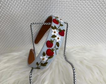 May include: A light blue headband with embroidered red strawberries and white flowers. The headband has a brown interior and is displayed on a silver metal stand. The design features green leaves and stems.