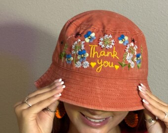 May include: An orange bucket hat with embroidered blue and white flowers and the words "Thank you" in yellow. The hat has a wide brim and is made of a soft fabric.