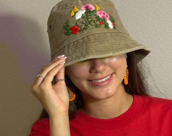 May include: A khaki-colored bucket hat with embroidered flowers in pink, white, yellow, and red. The hat is worn with a red t-shirt featuring a white floral design and orange beaded earrings.