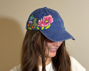 May include: A blue baseball cap with embroidered flowers in pink, yellow, blue, and orange. The floral design is on the left side of the cap. The cap has a curved brim and a classic six-panel design.