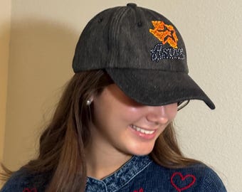 May include: A dark gray baseball cap with a curved brim. The cap features an orange and white embroidered design on the front. The design includes the word "Houston" in a stylized font. The cap is made of a textured fabric.