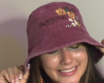 May include: A burgundy bucket hat with the word "Awesome" embroidered in black script. The hat features a floral design in shades of yellow, orange, and white. The hat is made of a soft, textured fabric.