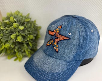 May include: Blue denim baseball cap with an embroidered star design. The star is orange with yellow and blue floral accents. The cap has a curved brim and a classic six-panel construction.
