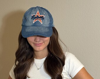 May include: A blue denim baseball cap with a star design. The star is outlined in orange and has the word "Astros" embroidered in the center. The cap is worn by a person wearing a white t-shirt.