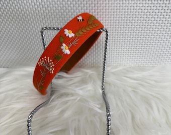 May include: An orange headband with embroidered white flowers, green leaves, and a small bee. The headband is displayed on a silver metal stand against a white, textured background. The embroidery details are visible.