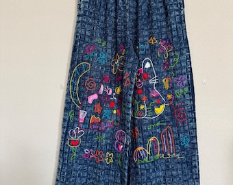 May include: Blue denim wide-leg pants with an elastic waistband. The pants feature colorful embroidered designs of flowers, a cat, and abstract shapes. The denim has a textured, grid-like pattern.