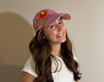 May include: A dusty rose baseball cap with embroidered floral designs. The cap features a large orange and yellow flower on the front, with smaller flowers and green leaves on the side. The cap has a curved brim and a classic six-panel design.