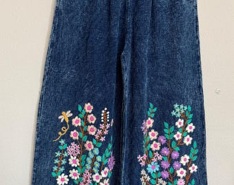May include: Blue denim wide-leg pants with an elastic waistband. The pants feature colorful floral embroidery along the lower legs, including pink, white, purple, and blue flowers with green leaves. A yellow dragonfly is embroidered on the left leg.