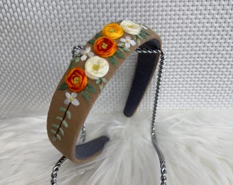May include: A brown headband with embroidered flowers in shades of orange, yellow, and white. The headband has a gray interior and is displayed on a silver metal stand. The floral design includes green leaves and small white flowers.