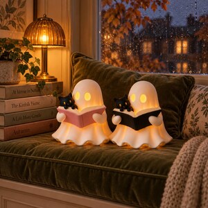 May include: Two white ghost-shaped lamps with glowing yellow eyes, each holding a small black cat figurine. One ghost holds an open pink book, while the other holds an open black book. The lamps are positioned on a green cushion near a stack of books.