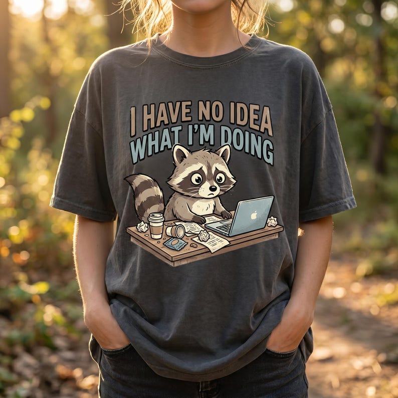 May include: Dark grey t-shirt with a cartoon raccoon at a desk with a laptop, coffee cup, and papers. The text "I HAVE NO IDEA WHAT I'M DOING" is printed above the raccoon. The shirt has short sleeves and a relaxed fit.