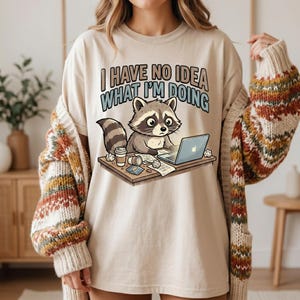 May include: Beige t-shirt featuring the text "I HAVE NO IDEA WHAT I'M DOING" and a cartoon raccoon using a laptop. The raccoon is at a desk with a coffee cup and crumpled papers. The shirt is being worn by a person.