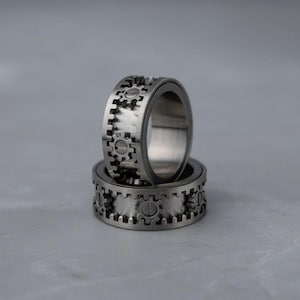 Puede incluir: Dos anillos plateados con diseño de engranajes. Los anillos están apilados, con el superior ligeramente inclinado. El diseño de engranajes está grabado alrededor de la banda exterior de cada anillo, creando una estética mecánica.