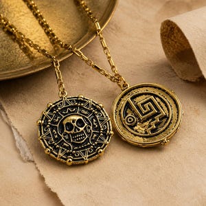 May include: Two gold-toned metal pendants on matching link chains. One pendant features a central skull surrounded by intricate radial patterns, while the other displays a geometric spiral design with a jagged line element in a circular frame.