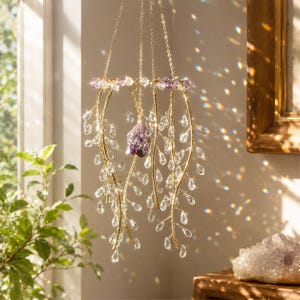 May include: A hanging mobile featuring a gold-toned circular frame adorned with purple beads and multiple dangling gold wire branches. Clear teardrop-shaped crystals hang from the branches, and a single raw purple amethyst crystal hangs in the center.