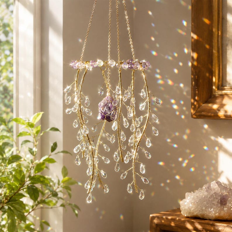 May include: A hanging mobile featuring a gold-toned circular frame adorned with purple beads and multiple dangling gold wire branches. Clear teardrop-shaped crystals hang from the branches, and a single raw purple amethyst crystal hangs in the center.