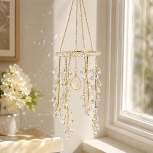 May include: A hanging mobile featuring a circular gold-toned frame suspended by thin chains. Numerous strands of gold wire hang from the frame, adorned with clear, teardrop-shaped crystal beads. A single, larger, irregular clear crystal hangs in the center.