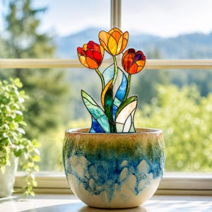May include: A stained glass tulip ornament featuring three flowers in red, orange, and yellow, with green and blue leaves. The ornament is placed inside a round ceramic pot with a gradient finish transitioning from cream at the base to blue and green.