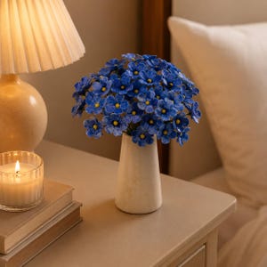 Op de afbeelding: Een boeket van kleine, levendig blauwe bloemen met gele harten, geschikt in een gladde, taps toelopende, gebroken witte keramische vaas. De bloemen hebben elk vijf bloemblaadjes en zijn samengevoegd tot een ronde, dichte bloemencompositie.