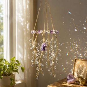 May include: A hanging mobile featuring gold-toned wire branches adorned with clear teardrop-shaped crystals. A central raw purple amethyst stone is suspended within the structure, which is topped by a ring of purple and clear crystal beads.