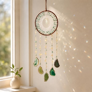 May include: A handcrafted dreamcatcher with a brown circular frame, adorned with green beads and hanging strands of gemstones. The dreamcatcher features a central web design and dangling agate slices in shades of green. It hangs in front of a window.