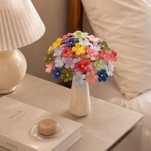May include: A bouquet of small, multi-coloured artificial flowers in shades of pink, blue, yellow, green, and purple, arranged in a smooth, light-coloured, tapered vase. The flowers feature five petals each with a small, contrasting yellow centre.