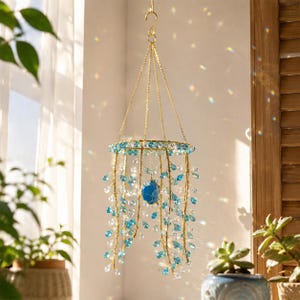 May include: A hanging mobile featuring a circular gold-toned metal frame with multiple strands of light blue and clear crystal beads. A single larger blue stone is suspended in the center. The structure is held by gold chains and a top hook.
