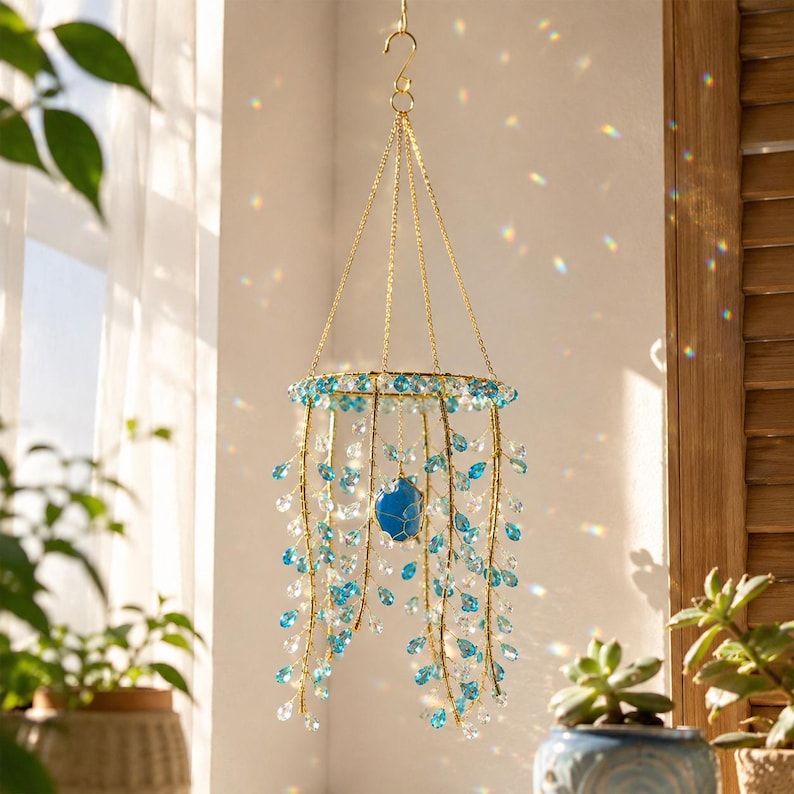 May include: A hanging mobile featuring a circular gold-toned metal frame with multiple strands of light blue and clear crystal beads. A single larger blue stone is suspended in the center. The structure is held by gold chains and a top hook.