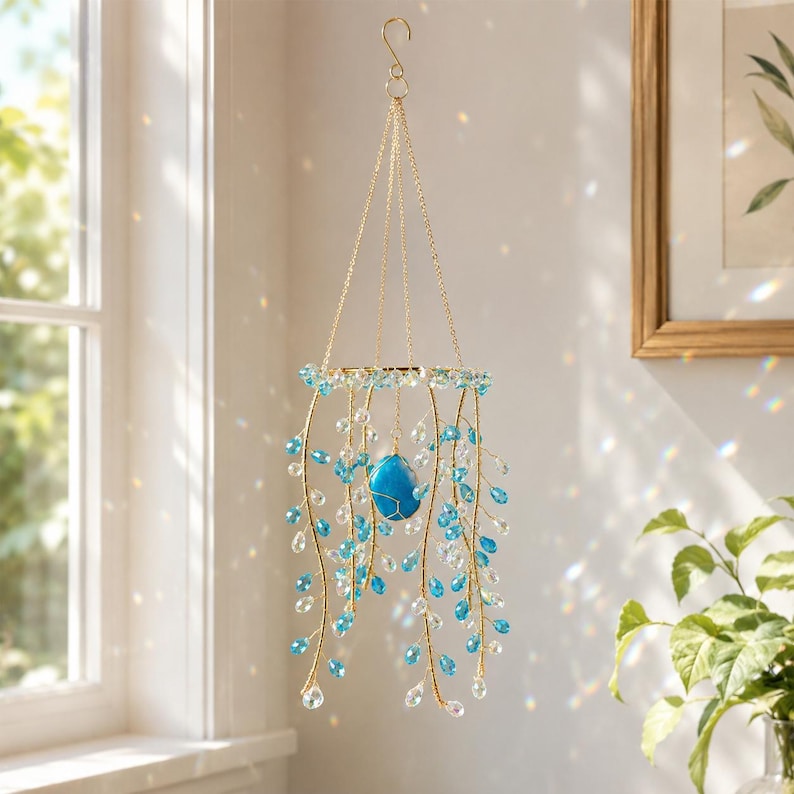 May include: A hanging mobile featuring a gold-toned circular frame with multiple gold wire strands adorned with clear and bright blue teardrop-shaped beads. A single, larger, smooth blue stone is suspended in the center of the mobile.