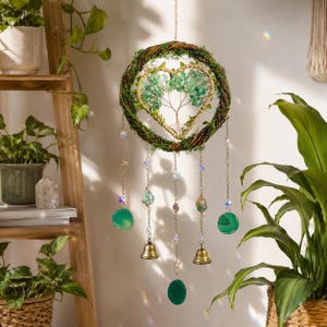 Può includere: Un ornamento da parete sospeso con una corona circolare avvolta nel muschio verde, che racchiude un albero della vita in filo metallico a forma di cuore con foglie di pietre preziose verdi. Cinque catenelle pendenti presentano cristalli trasparenti, fette di agata verde e due campanelli in ottone.